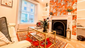 Uma sala de estar com sofá branco, mesa de centro de vidro, lareira com papel de parede floral e uma janela com persianas.