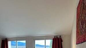 A bedroom with a large bed, two windows with curtains, a view of mountains, and a red tapestry on the wall.