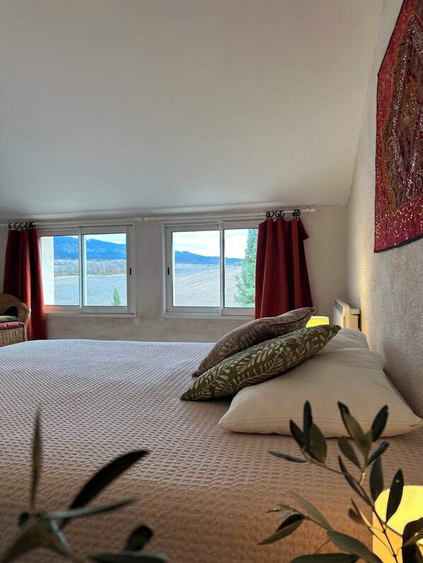 A bedroom with a large bed, two windows with curtains, a view of mountains, and a red tapestry on the wall.