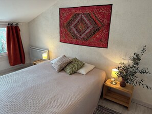 A neatly made bed with patterned bedding, a decorative wall hanging, a bedside lamp, and a potted plant.