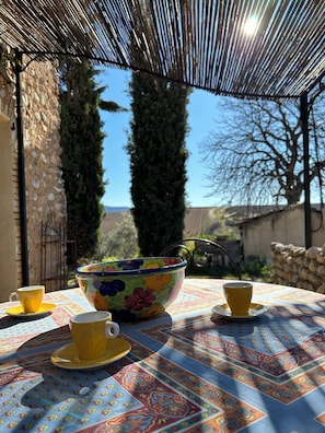 A table with a colorful tablecloth, a floral bowl, and two yellow cups on a patio with a stone wall and trees in the background.