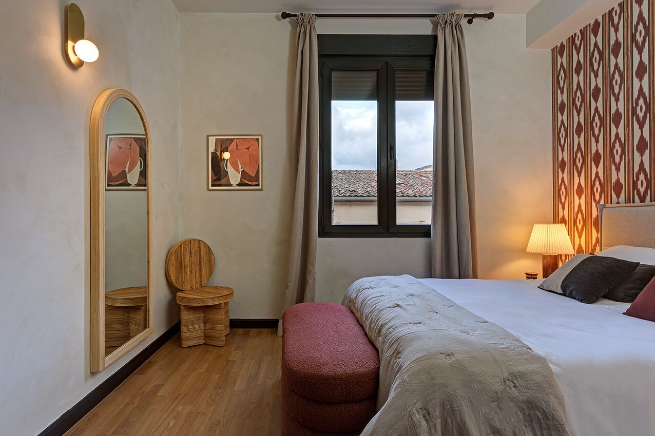 Una habitación de hotel con una cama, una silla de madera, un taburete pequeño, un espejo grande y una ventana con cortinas.
