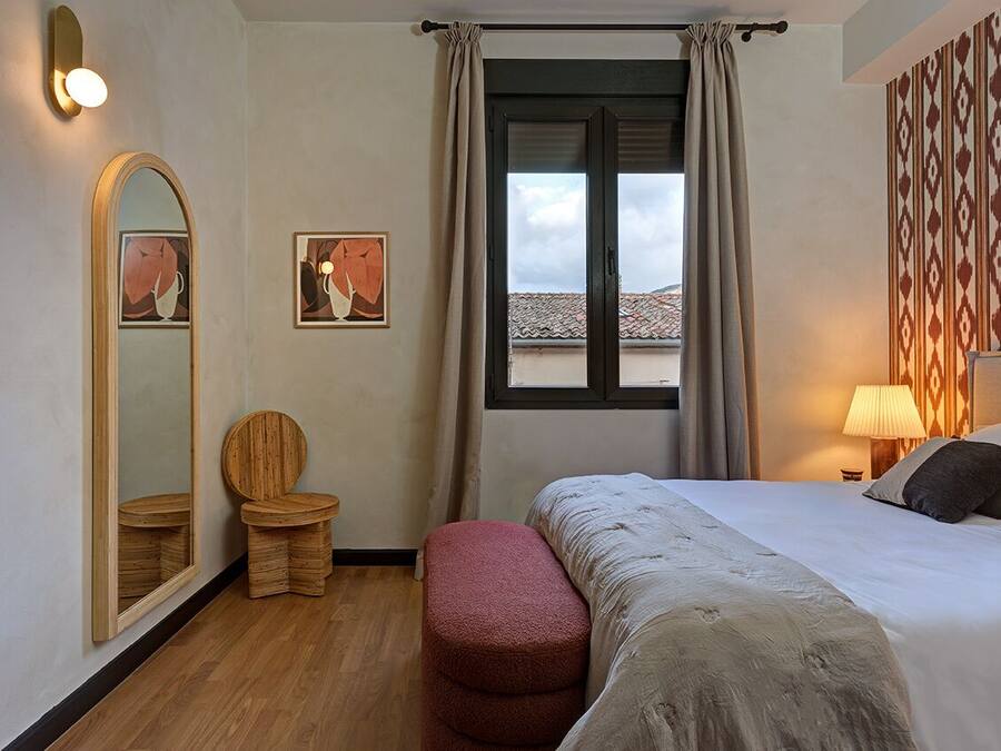 Una habitación de hotel con una cama, una silla de madera, un taburete pequeño, un espejo grande y una ventana con cortinas.