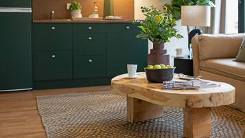 A modern kitchen with dark green cabinets, a wooden coffee table with a bowl of fruit, a white cup, and a magazine.