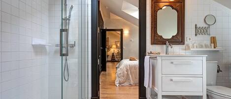 A modern bathroom with a glass shower enclosure, a white vanity with a mirror, and a view into a bedroom with a bed and a nightstand.
