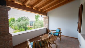 A balcony with a wooden ceiling, a brick wall, and a view of greenery. There are two wicker chairs and a small table. - Casa Nicole (Manerba del Garda)