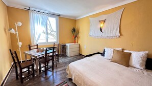 Una habitación con cama, un escritorio con sillas, una ventana con cortinas, una lámpara de pared y una planta pequeña.