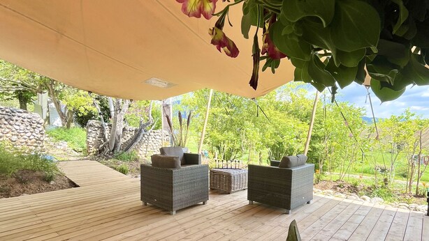 Un espacio al aire libre con piso de madera, mobiliario de mimbre y un toldo que ofrece sombra.