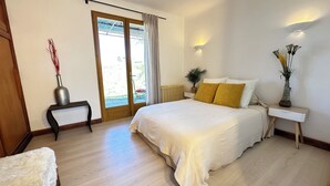 A bedroom with a large bed, a bedside table, a nightstand, a potted plant, and a view of the outdoors through a sliding door.