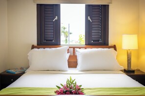 A hotel room with a bed, two white pillows, a nightstand with a lamp, and a window with shutters.