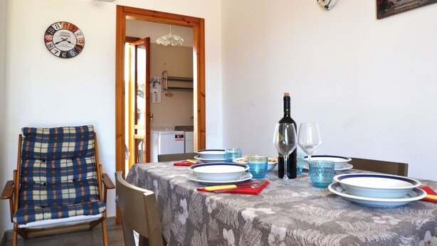 A dining area with a table set for two, a chair, a clock, and a kitchen visible through an open door. - Teoma Holiday Home With Climate and Parking for 5 Guests (Melendugno)
