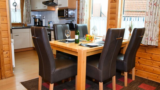 A wooden interior with a dining table set for two, surrounded by leather chairs. A kitchen with modern appliances is visible in the background. - House 47 the House to Stay for Holidays (Oberharz am Brocken)