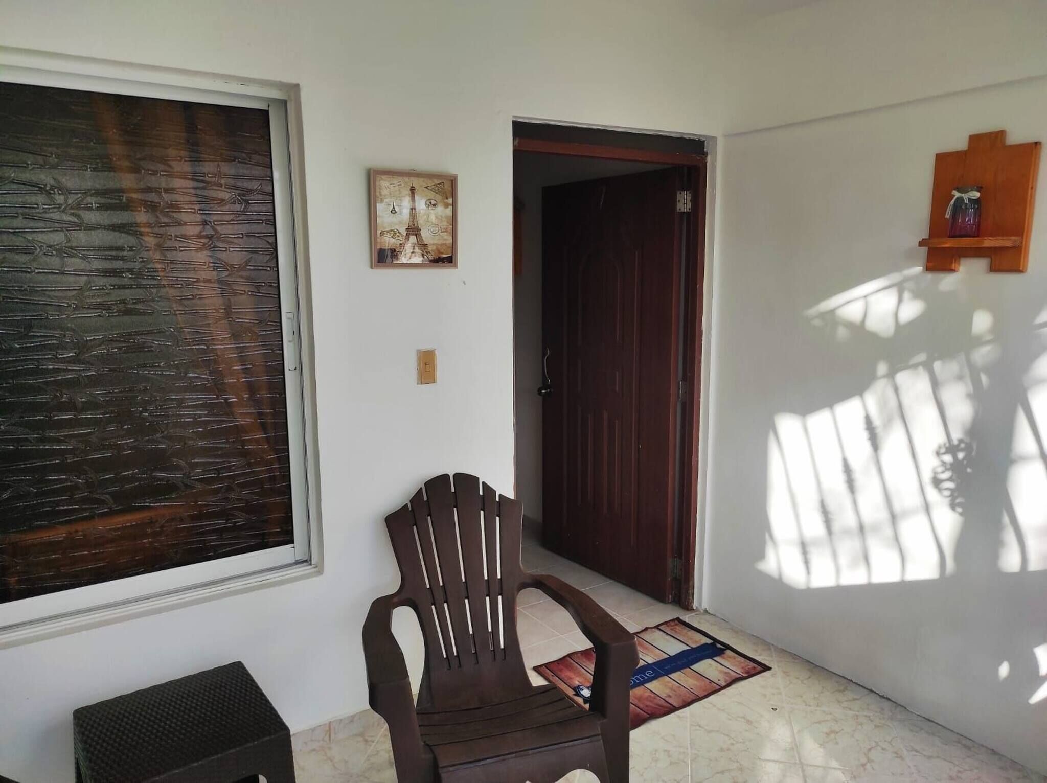 A room with a large window, a wooden door, a chair, and a small shelf with a jar.