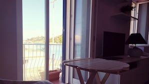 Quarto com mesa de madeira, televisão e varanda com vista para a praia e edifícios.