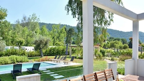 A poolside area with lounge chairs, a dining table with chairs, and a view of a lush garden and hills in the background. - Luxury Rental Villa for 14 People (Sithonia)