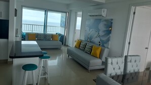 Un salon moderne avec un canapé gris, un coin repas avec une table noire et des tabourets blancs, et une grande fenêtre offrant une vue sur la mer.