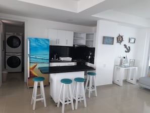 A compact kitchen with a central island, white cabinets, and a painting of a beach scene. There are four bar stools with teal seats. - 2 Bedroom Beachfront Apartment 2p1-al2 With Pool And Wifi (Cartagena)