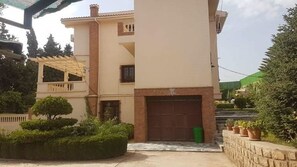 A two-story building with a garage, a covered porch, and a stone wall with potted plants. - Villa L'orangeraie, Wilaya de Tlemcen (Tlemcen)