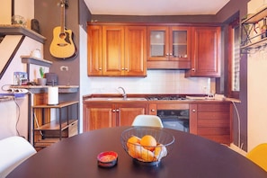 A kitchen with wooden cabinets, a dining table with a bowl of fruit, and a guitar on the wall. - - The Artist Village (Montoro)