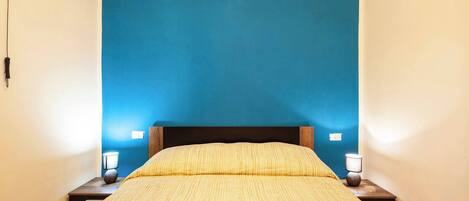 Un lit recouvert d’une couvre-lit jaune, avec une tête de lit sombre et deux lampes de chevet posées sur des tables de nuit en bois.