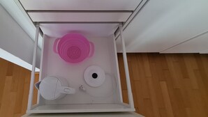 A white cabinet with a pink colander, a white kettle, and a white electric kettle inside. - Features of Good And Night (Neuss)