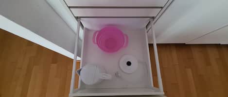 Un mueble blanco con un colador rosa, una tetera blanca y otra tetera eléctrica blanca en su interior.