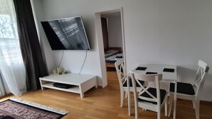 A room with a TV mounted on the wall, a bed with a wooden headboard, a white desk with a chair, and a small table with two chairs. - Features of Good And Night (Neuss)
