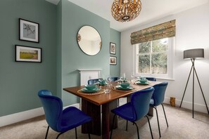 A dining room with a wooden table set for six, blue chairs, a large mirror, framed pictures on the wall, and a window with a view of greenery. - Beautiful Edwardian Home In Central Oxford (Oxford)