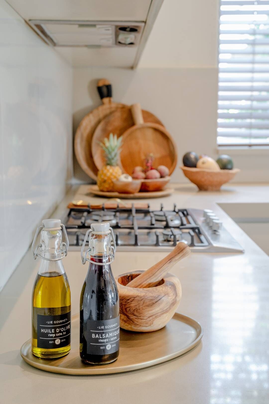 A kitchen counter with two bottles of olive oil, a wooden mortar and pestle, and a bowl of fruit.