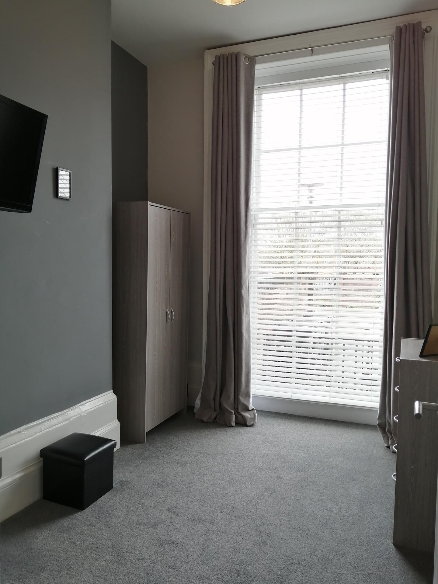 A room with a large window, a wardrobe, a flat-screen TV, and a small black ottoman.