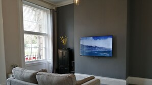 Ein Wohnzimmer mit grauem Sofa, einem flachbildfernseher an der Wand und einem Fenster mit Jalousien.