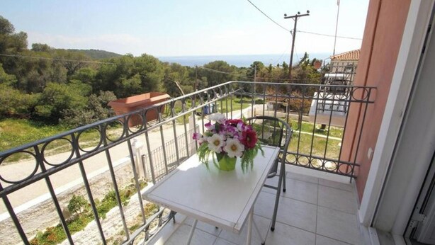 A balcony with a table and chairs, a flower vase, and a view of greenery and buildings. - Room-pelagos Studios by Frangki (Aegina)
