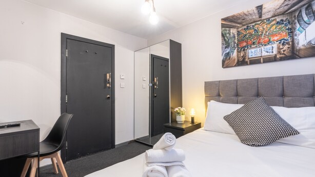 A hotel room with a bed, a desk, a chair, a wardrobe, and a painting on the wall. - Woodbury 2-4 (Lancaster)