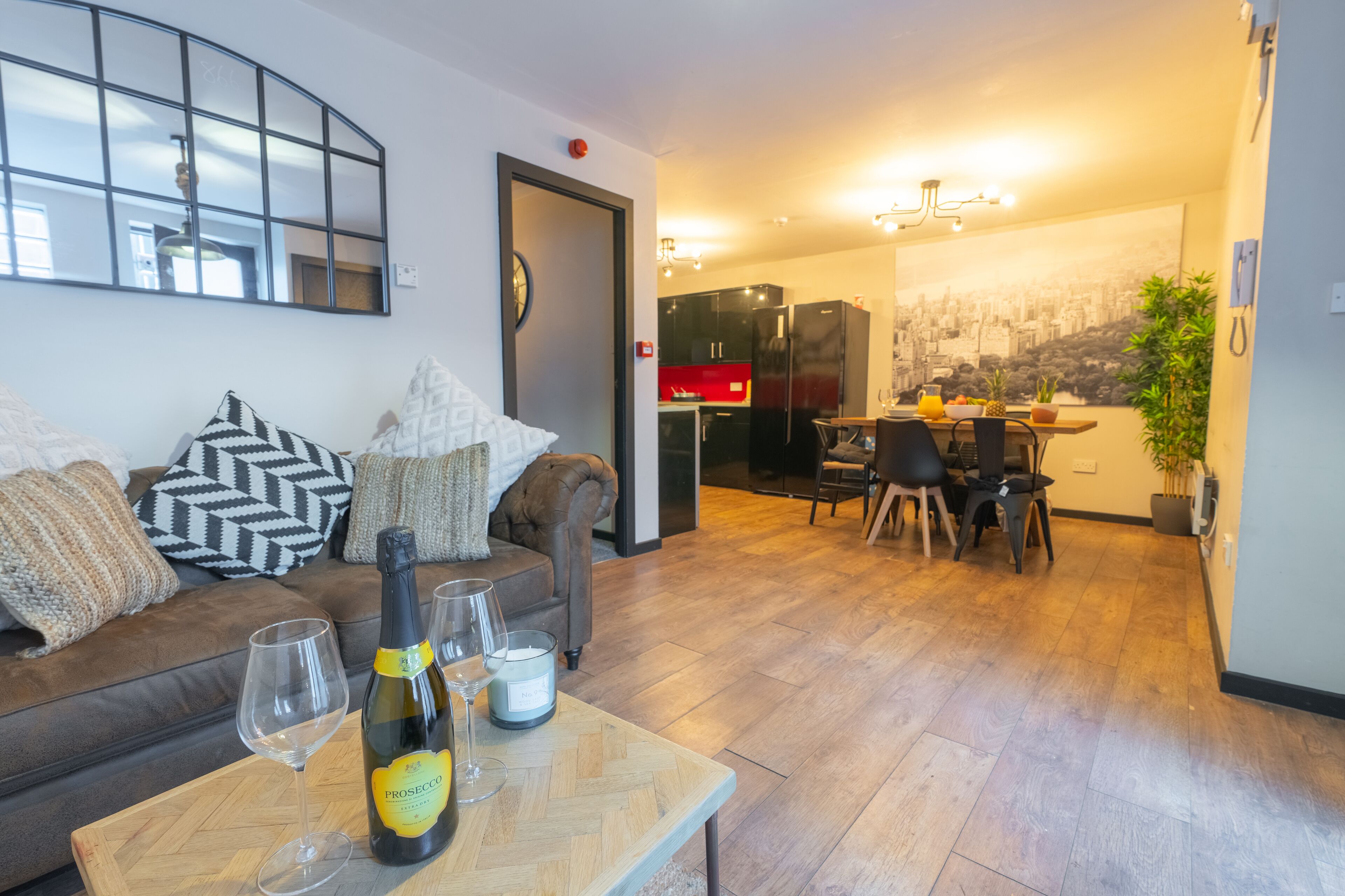 A living room with a sofa, a wooden coffee table with a bottle of Prosecco and glasses, and a dining area with a table and chairs.