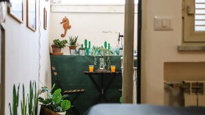 A narrow hallway with a green wall, a cactus-themed decoration, a small table with a vase and orange drink, and a wooden chair.