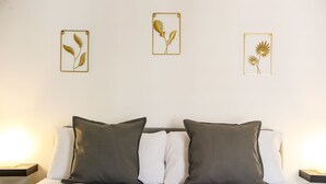 A bed with white bedding and two dark pillows, flanked by nightstands with lamps, and three framed botanical wall art pieces above.