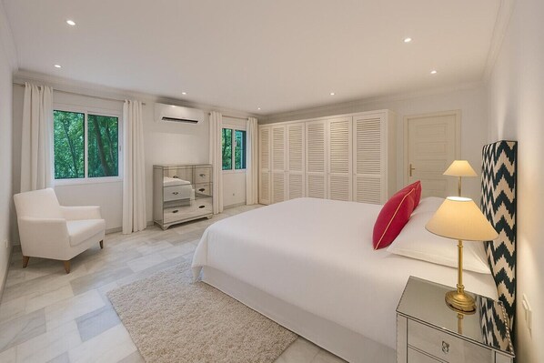 A bedroom with a large bed, a bedside table with lamps, a chair, a dresser, and a window with curtains.