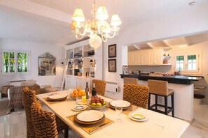 A modern kitchen with a dining area, featuring a chandelier, white cabinetry, and a view of the outdoors.