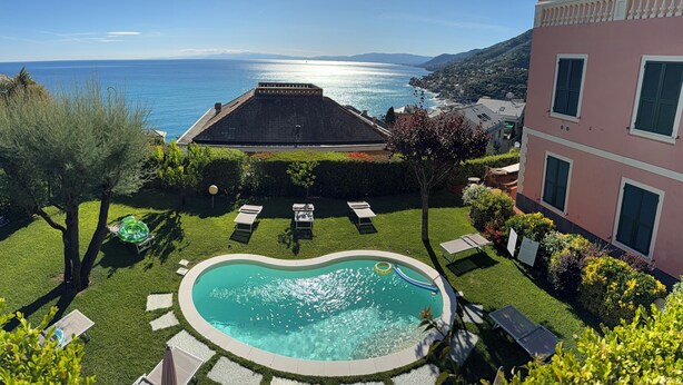 A swimming pool with a view of the sea, a building with balconies, and a well-maintained lawn.