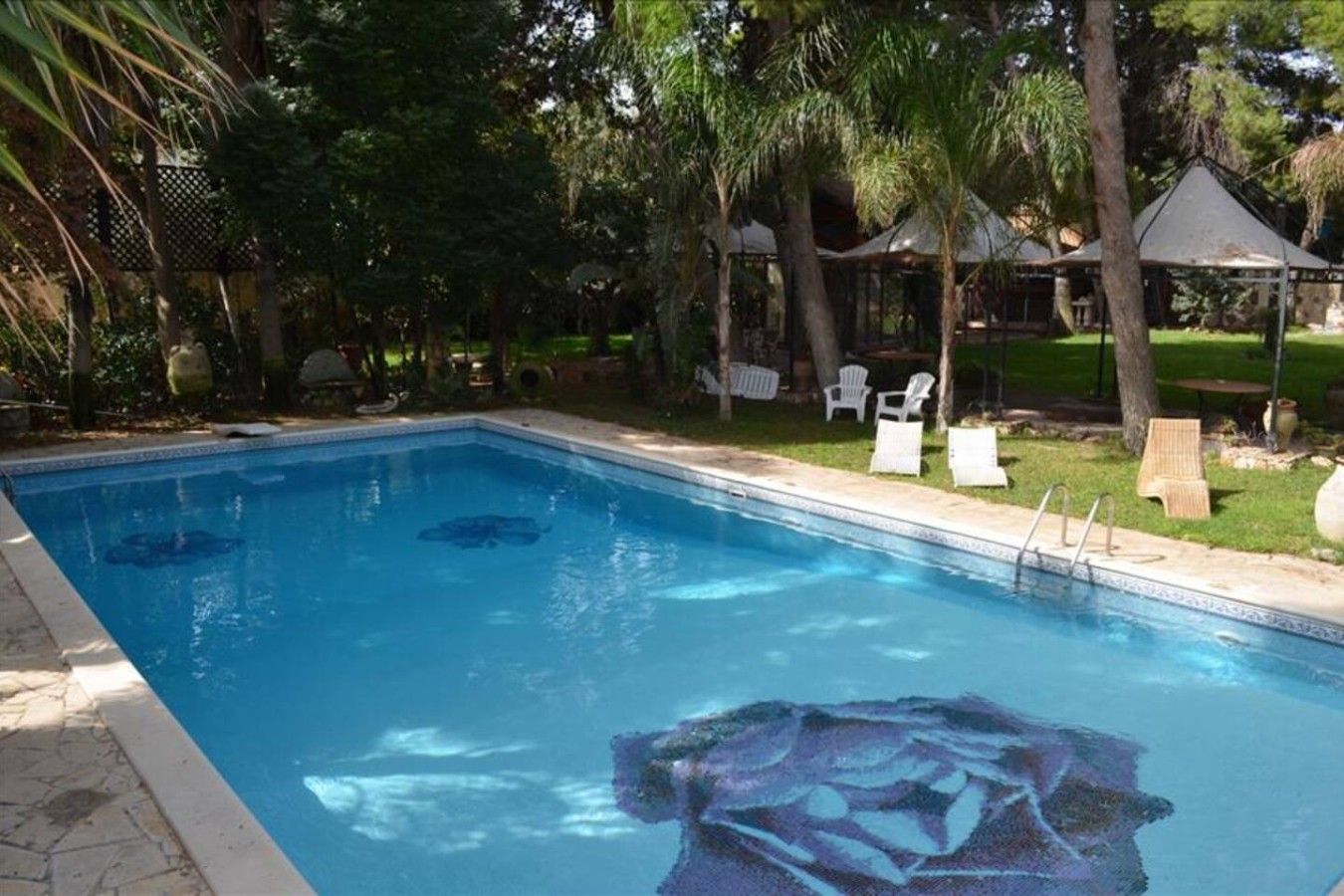 Ein rechteckiger Pool mit klarem, blauem Wasser, umgeben von einer gepflasterten Liegefläche. Im Hintergrund sind Liegestühle und eine Laube zu sehen.