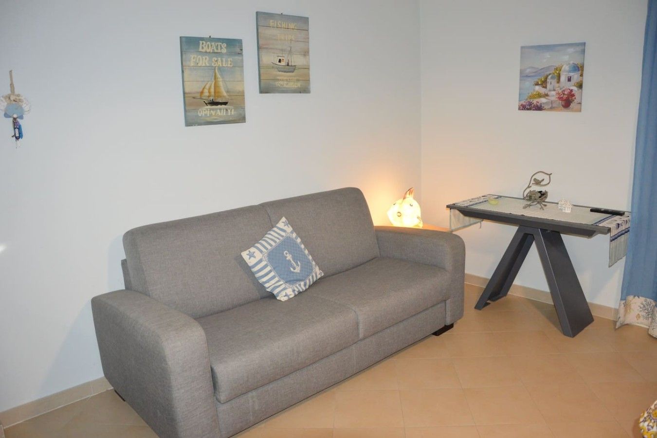 A grey sofa with a nautical-themed pillow, a small table with a lamp, and two posters on the wall.