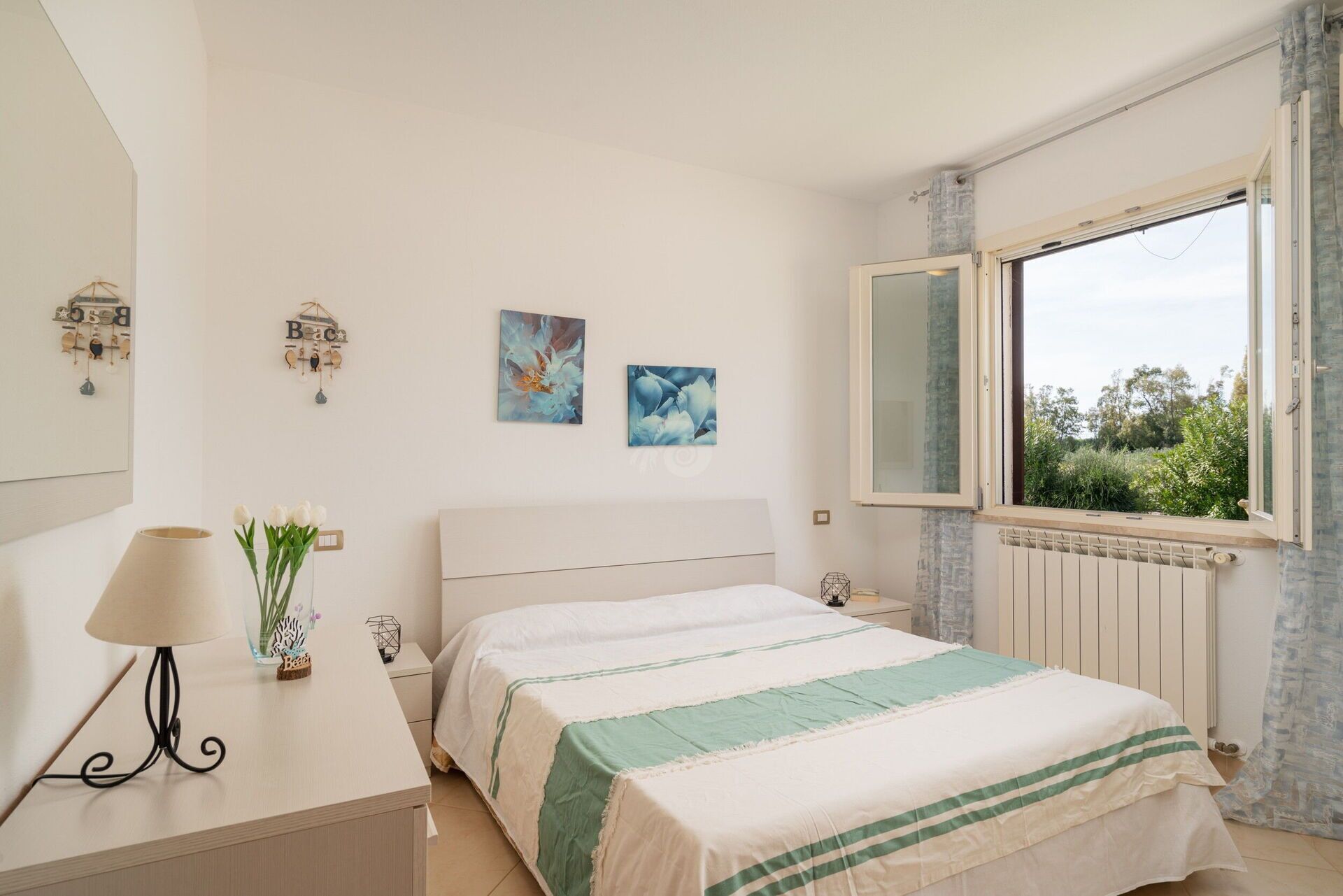 Un dormitorio con cama, mesitas de noche, lámpara, decoración en la pared y ventana con vistas.