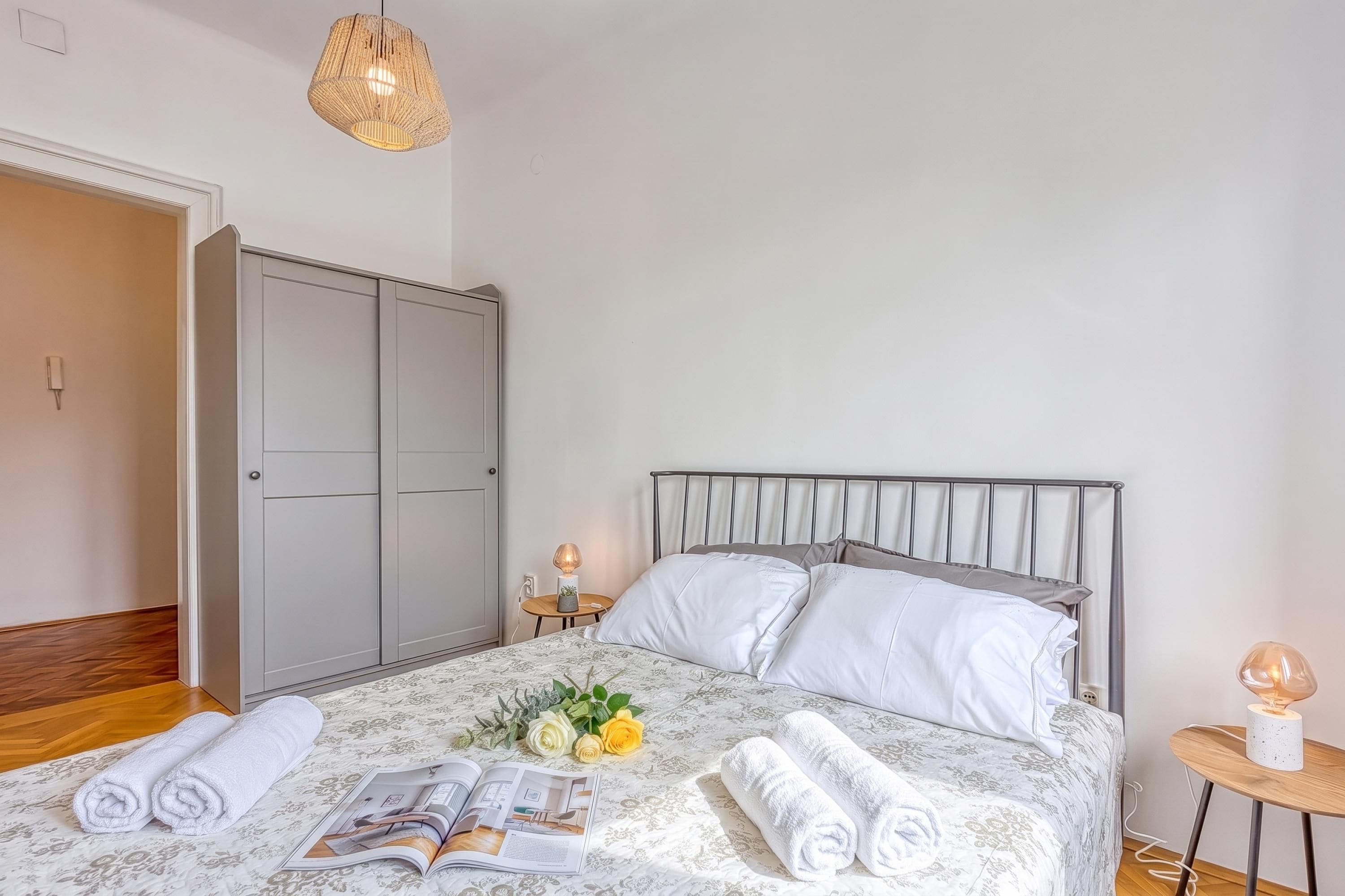 Una cama bien hecha con sábanas blancas, una colcha con estampado floral y una revista. Hay dos toallas enrolladas, una mesita con una lámpara y un aplique de pared.