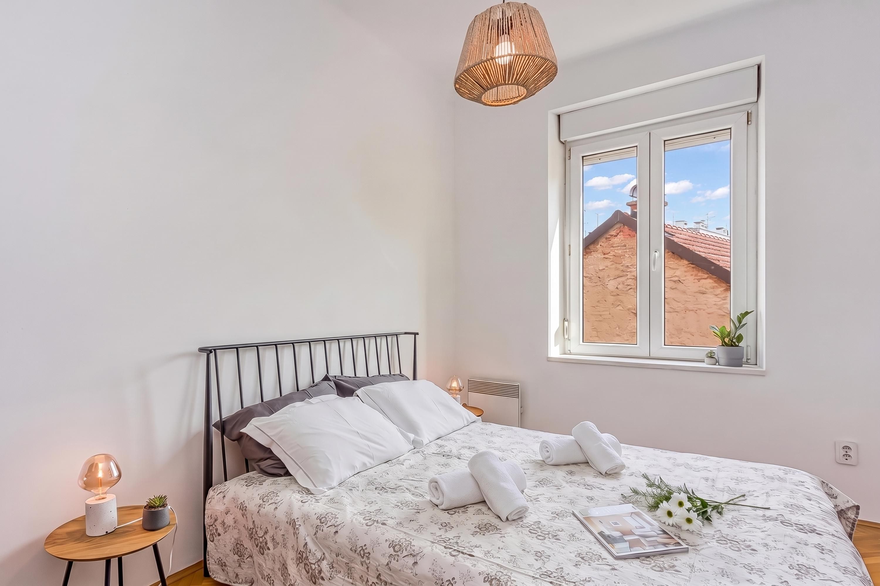 A bedroom with a bed, a bedside table, a lamp, a window with a view, and a hanging light fixture.