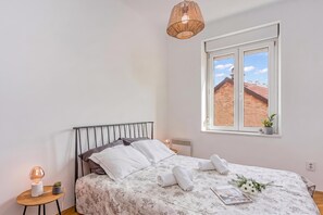 A bedroom with a bed, a bedside table, a lamp, a window with a view, and a hanging light fixture. - Spacious Apartment In Heart Of Zagreb (Zagreb)