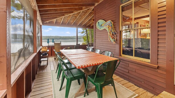 Een overdekt terras met een houten tafel en stoelen, uitzicht op het meer, en een bord dat gasten verwelkomt bij Camp Run-A-Muk.