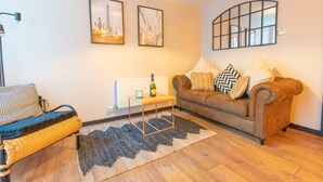 A living room with a brown sofa, a wicker chair, a small table with a bottle, and two framed pictures on the wall. - Woodbury 2-5 (Lancaster)
