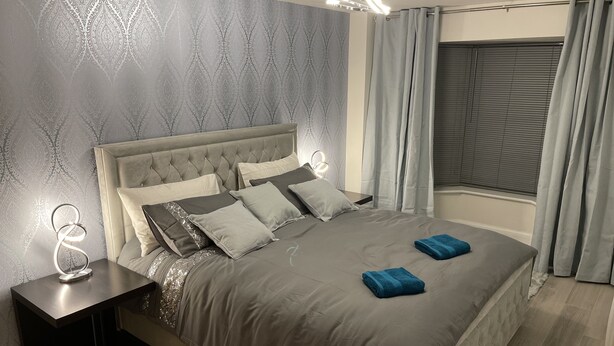 A bedroom with a tufted headboard, a bed with grey bedding, two bedside tables with lamps, and a window with curtains. - Lovely 1-bed Apartment in Town Centre Colchester (Colchester)