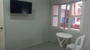 A small room with a flat-screen TV mounted on the wall, a window with a view of the outside, and a small white table with a chair.