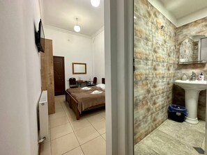 Una camera d'albergo con un letto, una scrivania, una sedia, uno specchio e un bagno con lavandino e doccia.